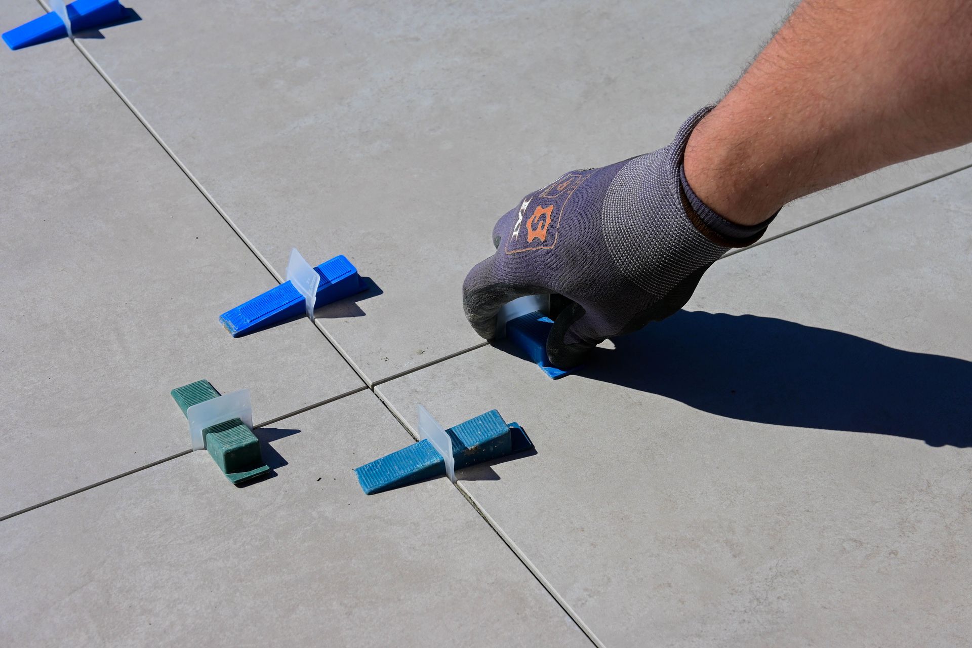 A close-up of a gloved hand installing tiles with spacers on a sidewalk.